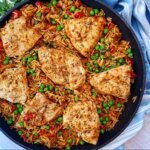 cast iron skillet placed on a light blue dish towel. It contains cooked chicken pieces in a bed of reddish brown rice with green peas and red bell peppers.